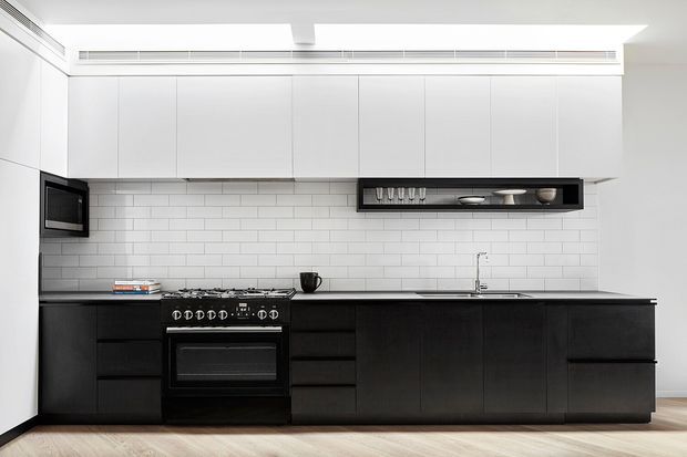 A bright, minimalist galley-style kitchen with a long run of matte black lower cabinetry, white handleless upper cabinets, and a white subway-tile splashback. Integrated appliances, a black freestanding range, stainless-steel sink tap, and a slim open display shelf complete the clean, monochromatic composition. Pale timber flooring and a linear ceiling vent add warmth and architectural detail.