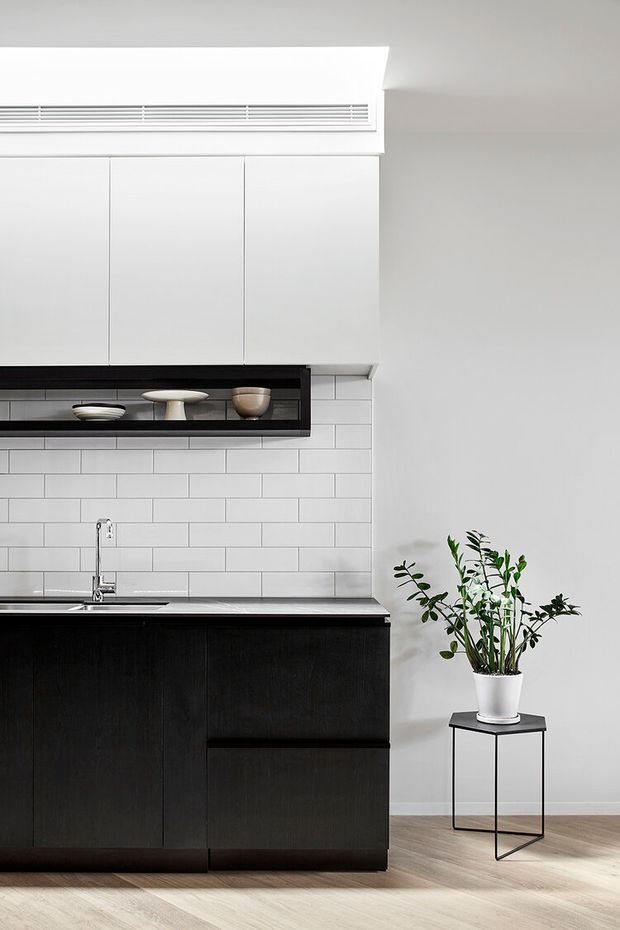 A minimalist kitchen with white upper cabinets, dark timber lower cabinetry, a slim dark benchtop, and a white rectangular subway-tile splashback. A black open display shelf holds ceramics, while a stainless-steel sink and tap sit beneath the cabinetry. Light timber flooring and a potted plant on a slender black stand add warmth to the bright, clean space.