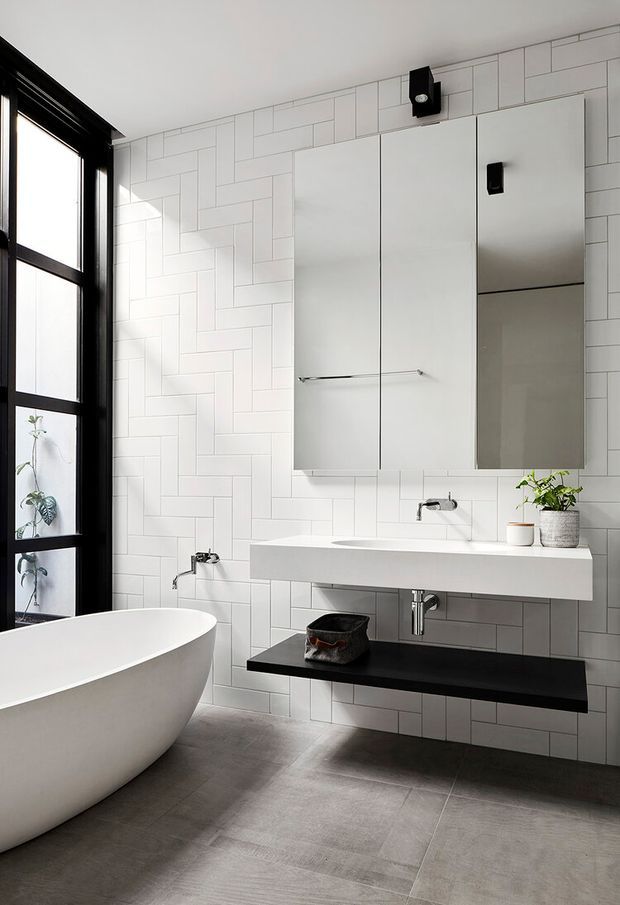 Contemporary bathroom with a freestanding white bathtub, wall-mounted double vanity, mirrored medicine cabinets, black floating shelf, black-framed floor-to-ceiling window, and white rectangular tiled walls in a geometric running-bond pattern. The room has grey floor tiles, black and chrome fixtures, and a clean monochrome palette with natural plants visible by the window.