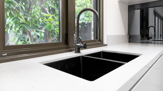 Close-up view of a contemporary kitchen work area featuring a large black undermount double sink, dark gooseneck faucet, and a bright white engineered-stone benchtop beneath brown-framed windows. White cabinetry and dark-framed glazing are visible, with leafy greenery outside.