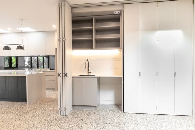 Bright, highly functional open-plan kitchen and concealed utility zone. The foreground is dominated by a large bank of tall white bi-fold cabinet doors, with an integrated sink and workbench behind them, open grey shelving, and warm under-shelf lighting. The adjoining kitchen has a dark island, light cabinetry, pendant lights, and windows overlooking greenery. Terrazzo-style flooring runs throughout.