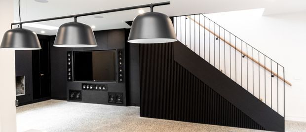 A contemporary dark-toned media room viewed from the living area, featuring a large built-in home theatre wall with screen and speakers, three oversized black pendant lights, and a dramatic black stair balustrade with vertical metal rods and timber handrail. Light carpet and white walls provide contrast.