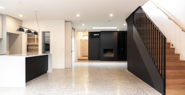 Open-plan contemporary residential interior featuring a white kitchen with a dark island, pendant lighting, built-in shelving, a black feature wall with fireplace, polished terrazzo flooring, and a timber staircase enclosed by a dark vertical-slat balustrade.