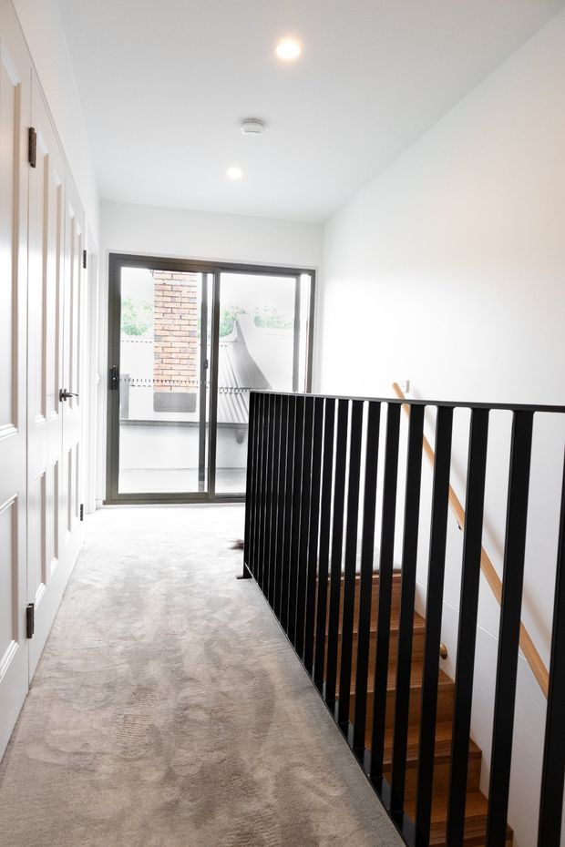 Bright contemporary upstairs landing or hallway with pale carpet, white walls and ceiling, paneled interior doors, and a prominent black vertical-baluster stair balustrade. A glazed sliding door at the end opens to an outdoor balcony or terrace, while the stair opening reveals timber stair treads and a timber handrail.