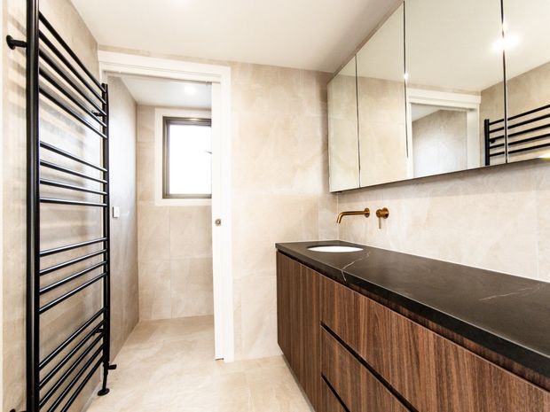 Contemporary bathroom with a floating dark timber vanity, black stone benchtop, integrated basin, wall-mounted brass tapware, large mirrored cabinetry, beige stone-look wall and floor tiles, and a black ladder-style heated towel rail. A small enclosed shower or wet-area space is visible through the doorway.