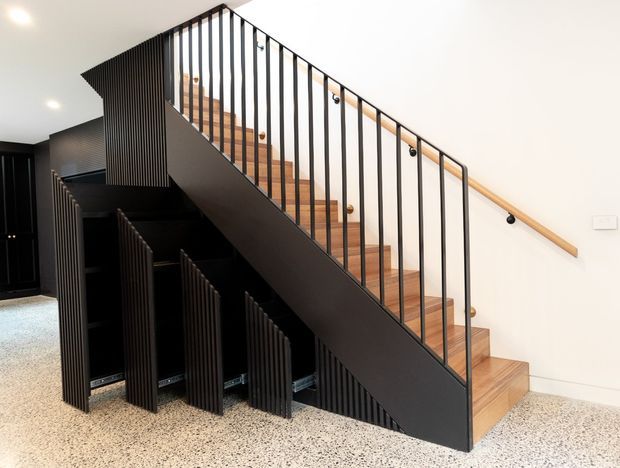 Interior view of a contemporary stairway with warm timber treads, slim black metal balustrades, and a black sculptural stair stringer. The staircase incorporates ribbed dark storage or screening elements beneath it, set against white walls and a terrazzo-like floor.