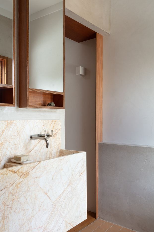A refined bathroom vanity area with a freestanding stone-clad basin, wall-mounted brushed metal tapware, integrated marble-look splashback, timber-framed mirrored cabinetry, warm timber ceiling detail, plastered walls, and terracotta-toned tiled flooring. The room has a warm, earthy and minimalist character with strong natural stone veining and timber accents.