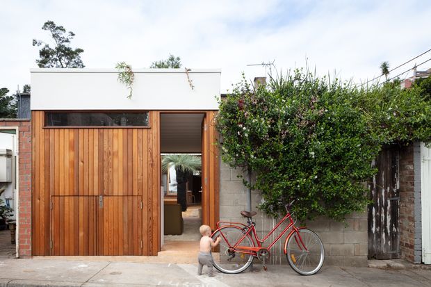 Street-facing view of a compact contemporary residential extension or dwelling with a tall vertical timber garage or entry door, white flat-roofed parapet, narrow horizontal window, exposed brick side wall, and a recessed doorway opening toward an interior space. Dense planting and a red bicycle sit in front of the facade.