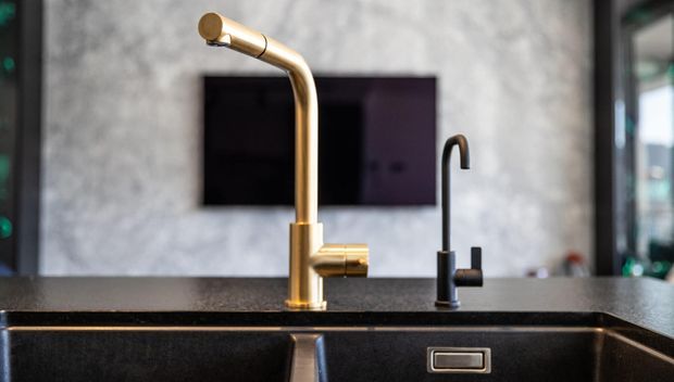 Close-up of a modern kitchen sink area with a dark stone or composite benchtop, integrated dark sink, and two contrasting mixer taps: a prominent brushed brass gooseneck tap and a slimmer black tap behind it. A textured grey wall and dark cabinetry or appliances are visible in the background.