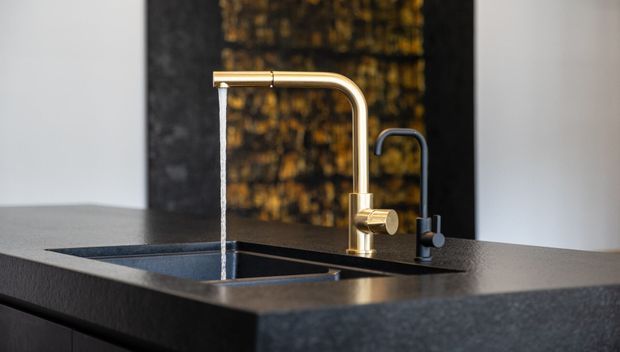 Close-up of a modern kitchen sink set into a dark stone benchtop, featuring a brushed brass gooseneck tap with a separate dark secondary faucet. The scene has a refined contemporary look with a dark textured feature wall and neutral surrounding surfaces.