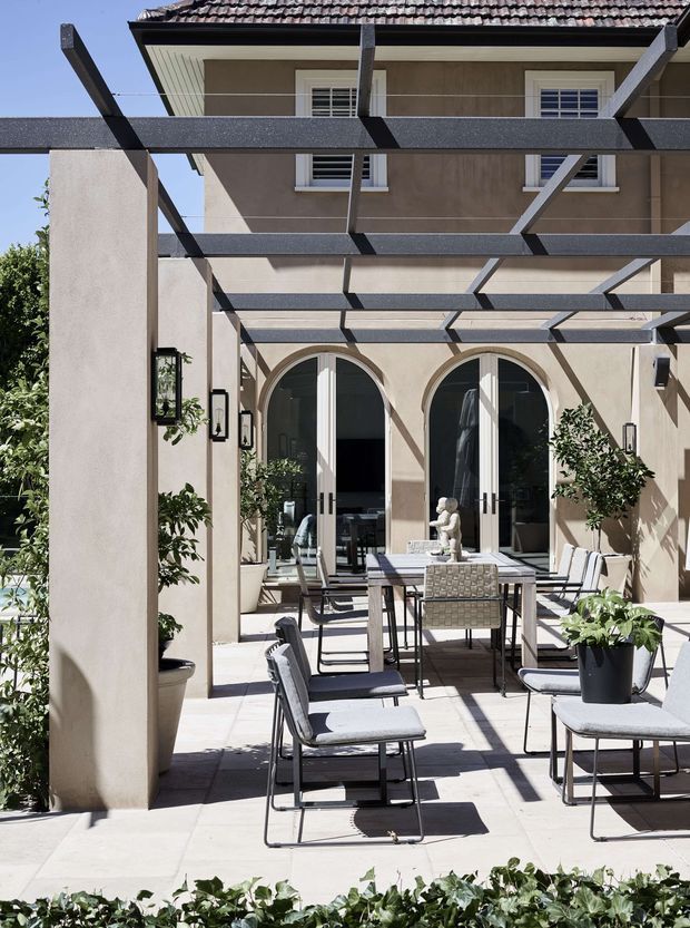 A bright, elegant outdoor terrace courtyard enclosed by stucco walls and a dark steel pergola. The paved patio is furnished with a dining table, upholstered dining chairs, potted plants, and decorative objects. Arched French doors open into the building, while the upper level features rendered walls, white-framed windows, and a tiled pitched roof. Strong sunlight creates geometric pergola shadows across the terrace.