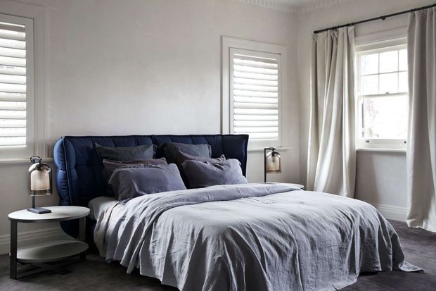 A refined bedroom with a deep navy upholstered bed, layered grey-blue bedding, and matching bedside tables with lamps. White walls feature decorative crown moulding, plantation shutters, and a large curtained sash-style window. The room has a calm, cool-toned palette and traditional detailing.