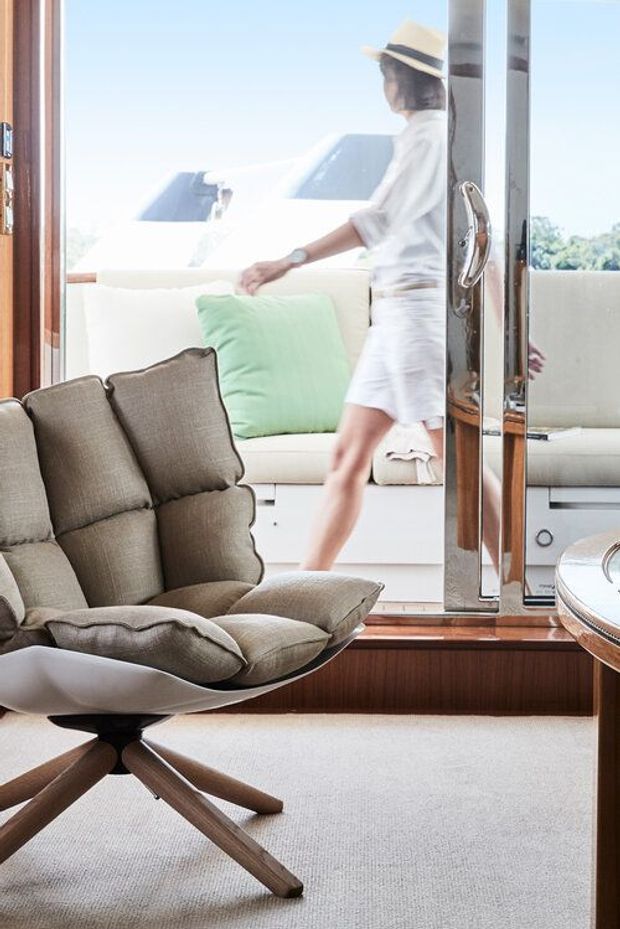 Bright, refined interior corner featuring a sculptural upholstered lounge chair with timber legs, pale carpet, and dark timber-framed full-height glazing or doors. A mirrored or reflective glazed panel and a white outdoor lounge with mint cushion are visible beyond, creating an airy connection to the exterior. The image has a soft neutral palette and abundant natural light.