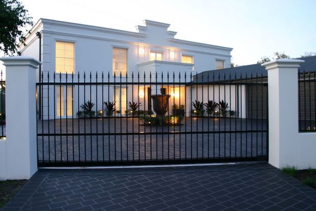 A grand white rendered multi-storey residence viewed from the street at dusk, enclosed by a black spear-top metal security fence and matching sliding driveway gate. The formal facade features tall illuminated windows, classical cornices and parapets, white masonry gateposts, a dark tiled driveway, and a landscaped courtyard with a central urn planter.