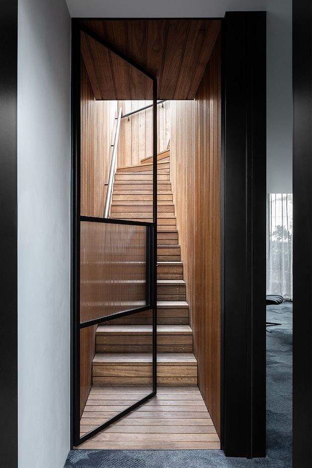 A narrow, enclosed timber-lined stairway viewed through a black-framed glazed door. Warm horizontal and vertical wood panelling continues across the treads, walls and ceiling, with a simple metal handrail. The stairwell opens toward a darker carpeted interior room at right, creating a strong contrast between the warm timber stair enclosure and the surrounding dark framing.