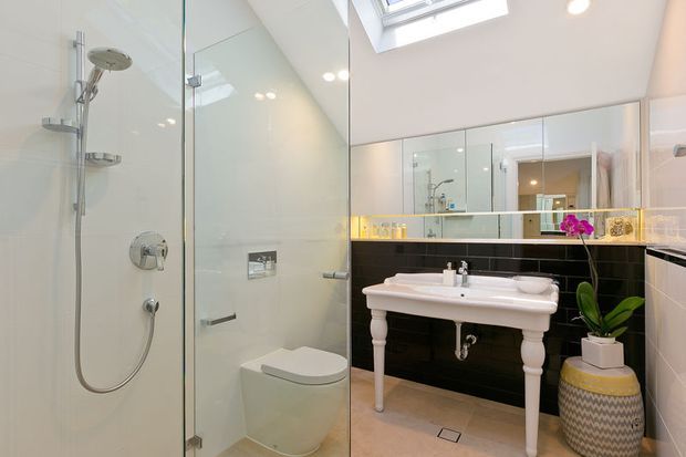 Bright, refined bathroom with a frameless glass shower enclosure, wall-hung toilet, freestanding white basin vanity with turned legs, large mirrored cabinetry, black glossy tiled feature wall, pale tiled surfaces, and a skylight. A potted orchid adds a small decorative accent.