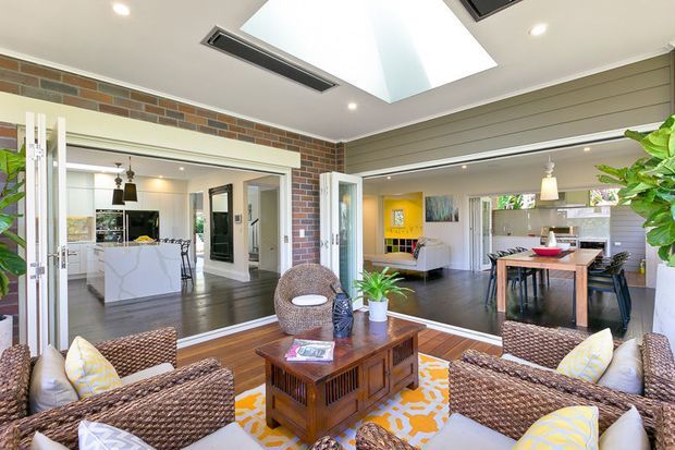 Bright open-plan living and dining area with a woven rattan seating arrangement in the foreground, centred around a timber coffee table and patterned rug. Large glazed openings connect the room to a modern white kitchen and adjacent dining space. Exposed brick walls, horizontal light-coloured wall cladding, dark timber flooring, skylight, and abundant indoor plants create a warm, airy interior.