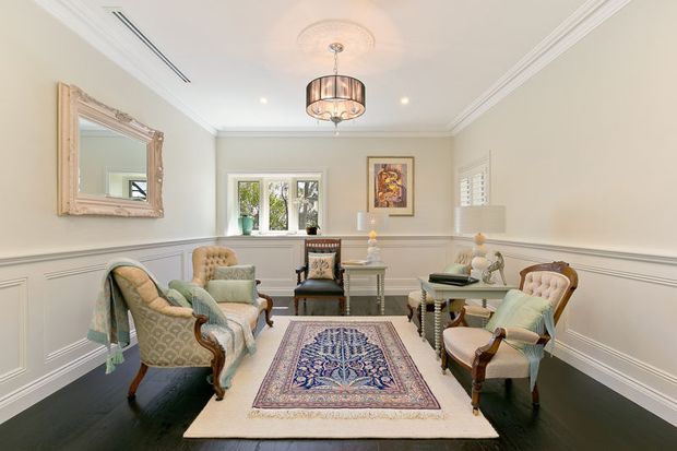 A refined sitting room with pale paneled walls, ornate crown moulding, dark timber flooring, and traditional upholstered armchairs arranged around layered rugs. A decorative chandelier, framed artwork, carved mirror, side tables, and plantation shutters create a classic, elegant interior.