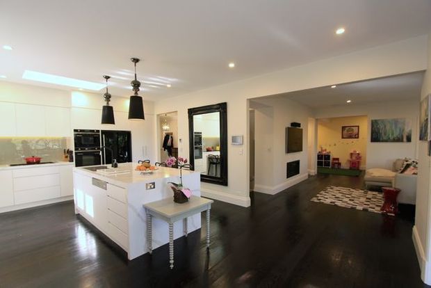 Bright open-plan residential interior featuring a white kitchen with a central island, integrated appliances, pendant lighting, dark timber flooring, and a connected living area with a fireplace, television, sofa, and rug. A large framed mirror and light neutral walls contribute to the spacious, polished appearance.