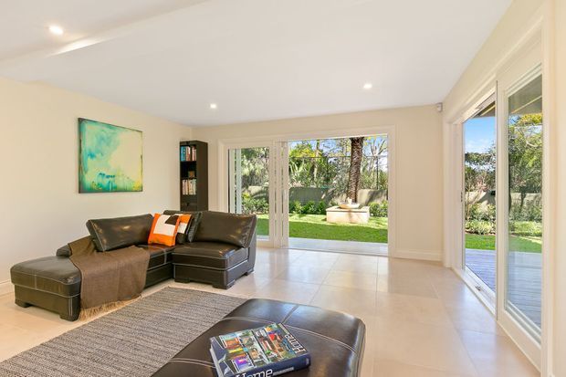 Bright, contemporary living room with a dark leather sectional and ottoman, neutral tiled flooring, pale cream walls, recessed ceiling lights, and large glazed doors opening to a landscaped garden. A colorful abstract artwork and a textured rug add accents, while the generous glazing creates an airy indoor-outdoor connection.