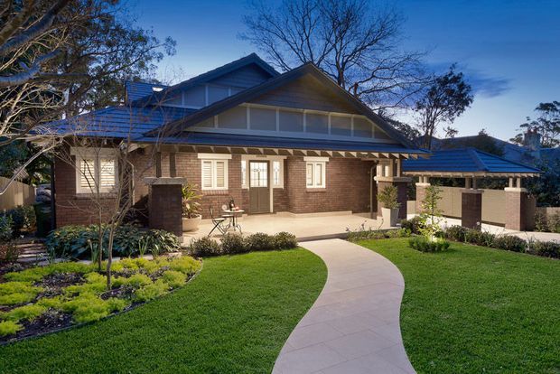 Street-facing view of a substantial single-storey brick home with a prominent gabled roof, broad covered entry porch, white-framed windows, and an attached side carport or covered parking area. A curved concrete walkway leads through a manicured front garden and lawn.