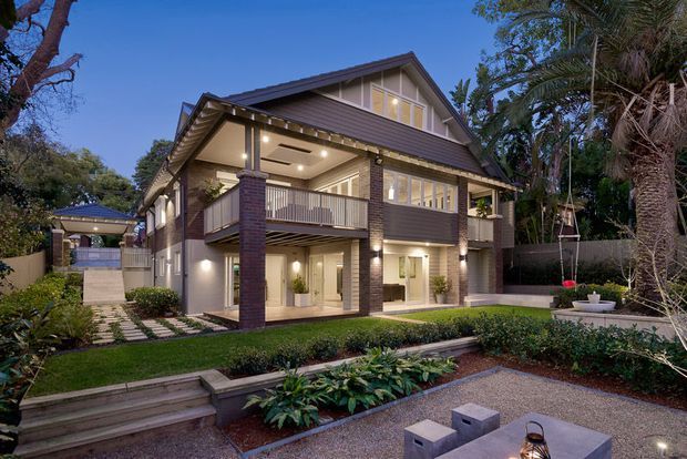 A large two-storey residential house photographed at dusk, featuring a steep gabled roof, dark brick and rendered walls, extensive white-framed glazing, upper-level balconies, deep eaves, and warm integrated exterior lighting. The house is set within a landscaped garden with paved paths, raised planting beds, and a small outdoor seating area.