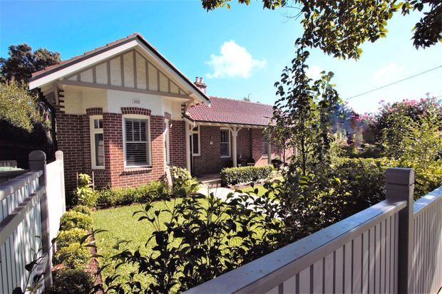 A single-storey red-brick house viewed from a landscaped front garden behind a white picket-style fence. The home has a gabled roof with decorative timber gable detailing, white-framed windows and doors, and a tiled roof over the main section. Mature shrubs and trees surround the sunny residential facade.