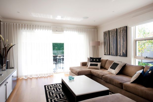 Bright, spacious contemporary living room with a large sectional sofa, white rectangular coffee table, patterned area rug, sheer floor-to-ceiling curtains, timber flooring, and framed abstract wall art. Glazed doors open toward an outdoor deck, while a large window brings in natural light.