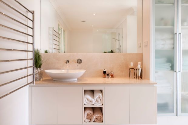 Bright contemporary bathroom vanity with a floating white cabinet, integrated pale stone benchtop and backsplash, vessel basin, large mirror, chrome heated towel rail, glass shower enclosure, and frosted-glass storage cabinetry. Soft neutral finishes create a clean, refined feel.