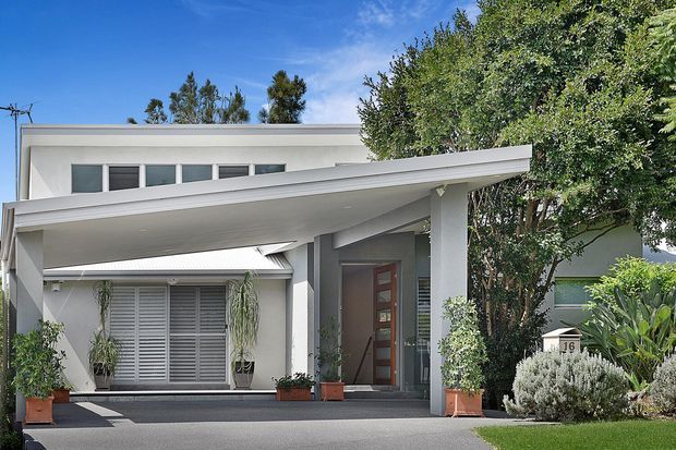 Street-facing view of a contemporary two-storey residence with a broad skillion-style porch roof and covered carport or entry canopy. The white rendered facade features horizontal clerestory windows, a timber-panelled entry door, louvred front glazing, square columns, and landscaped planting around the paved approach.