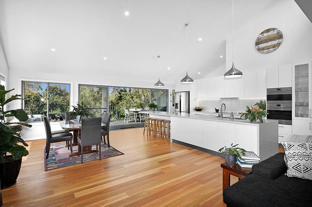 Bright open-plan contemporary kitchen and dining area with white handleless cabinetry, a large island with an integrated sink, stainless-steel appliances, pendant lights, timber flooring, and expansive windows opening toward a leafy outdoor setting. A dining table and living seating are also visible.