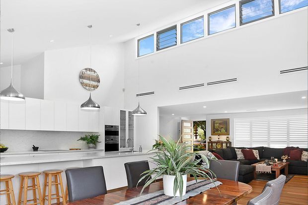 Bright open-plan kitchen, dining and living area with a high ceiling, clerestory windows, white cabinetry, a long island with timber stools, pendant lights, and a timber dining table. The adjoining living zone includes a dark sectional sofa, plantation shutters and indoor greenery. The room has a clean, contemporary feel with abundant natural light.
