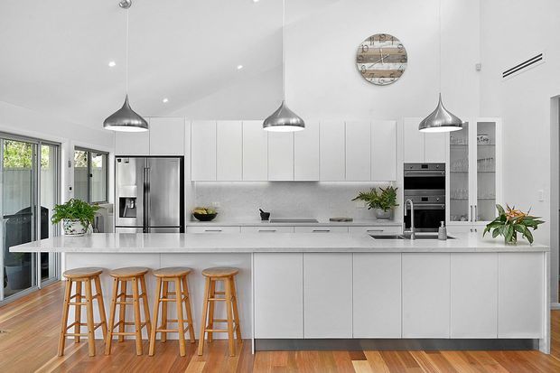 Bright contemporary white kitchen with a large central island, four timber bar stools, white handleless cabinetry, integrated stainless-steel appliances, and three suspended metallic pendant lights. Full-height glazing and a glass door bring in natural light, while warm timber flooring adds contrast.