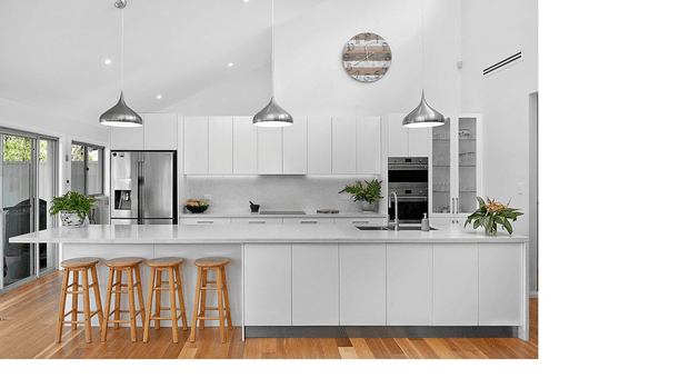 Bright, spacious contemporary white kitchen with a large waterfall-edge island, four timber bar stools, integrated stainless-steel appliances, extensive white cabinetry, pendant lighting, and warm timber flooring. Glazed doors and windows provide natural light, while plants and a round wall feature add subtle visual interest.