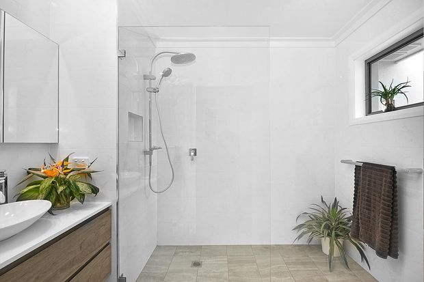 Bright contemporary bathroom featuring a walk-in glass shower with rainfall and handheld fixtures, a floating timber-look vanity with a white vessel basin, large pale wall tiles, light floor tiles, a horizontal window, towel rail, and indoor plants.