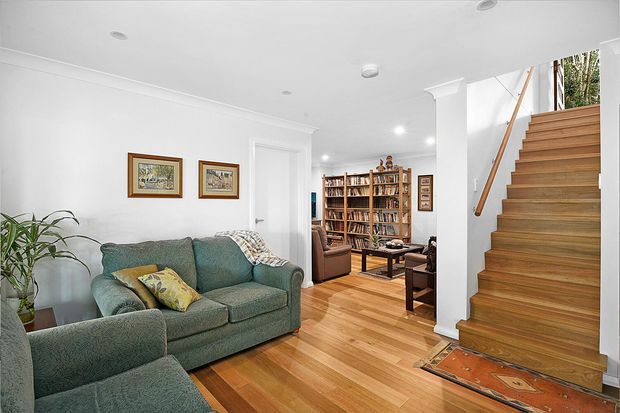 Bright, traditional living area with green upholstered sofas, timber flooring, framed artwork, and a substantial built-in bookcase visible beyond. A timber staircase with a simple wooden handrail rises along the right side, creating a connection to the upper level. White walls and ceiling provide a clean backdrop, while a patterned runner adds warmth at the stair base.