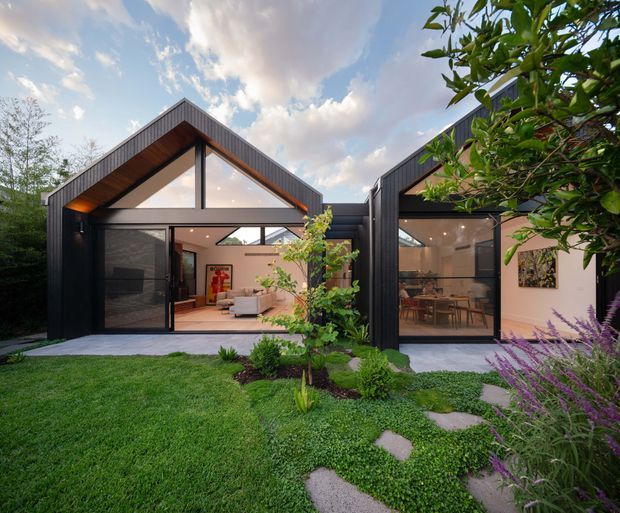 A contemporary single-storey residential extension or pavilion composed of two dark-clad gabled volumes with extensive floor-to-ceiling glazing. The open living and dining spaces are visible through the glass, connected to a landscaped garden with stepping stones, lawn, planted beds, and mature greenery. The architecture features strong black framing, exposed timber-toned soffits, triangular clerestory glazing, and a sheltered outdoor threshold.