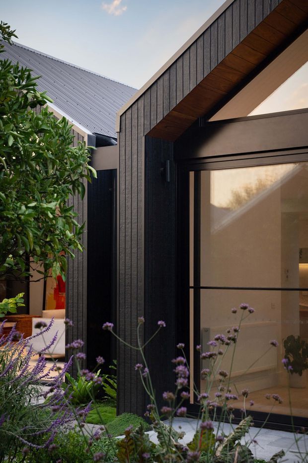 A close-up exterior view of a contemporary courtyard-facing building, featuring dark vertically textured cladding, a sharply pitched gable roof with warm timber soffit lining, large glazed doors or windows in dark frames, and a landscaped garden with purple flowering plants in the foreground. A secondary light grey metal-roofed structure is visible behind the main facade.