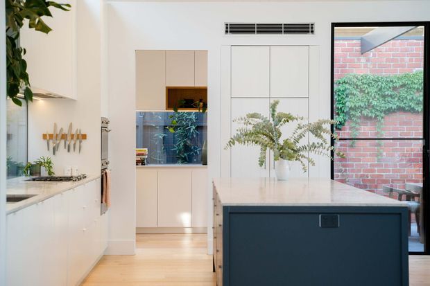 Bright contemporary kitchen viewed from the main cooking area, featuring a large dark island with a pale stone benchtop, white cabinetry, pale timber flooring, integrated appliances, and black-framed glazing opening to a brick courtyard. A built-in pantry or storage wall is visible beyond the island, with greenery adding a natural touch.