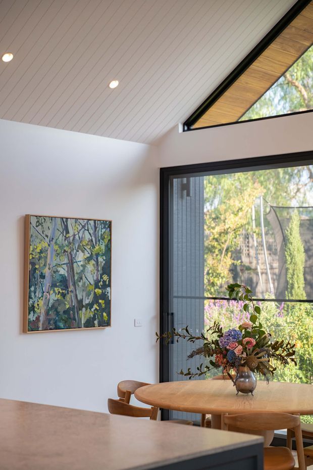 Bright contemporary dining area adjoining a kitchen, with a round timber dining table and chairs beside large black-framed glazed doors. The room features a white painted vertical-panel ceiling, white walls, a warm timber-lined triangular clerestory window, and a colourful floral artwork. Garden greenery is visible through the glazing.