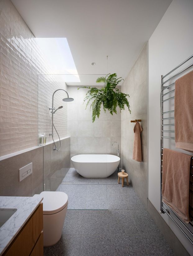 Bright contemporary bathroom with a freestanding white bathtub, walk-in shower behind a glass screen, wall-mounted rainfall shower, terrazzo-style flooring, pale textured wall tiles, timber vanity, and a chrome heated towel rail with blush towels. A skylight and hanging fern add natural light and greenery.
