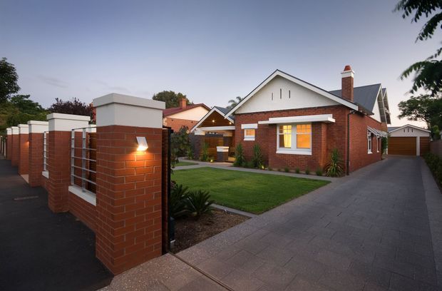 Exterior view of a suburban brick residence at dusk, featuring a gabled roof, white rendered gable ends, red brick walls, a paved driveway, landscaped front lawn, brick boundary pillars, and a detached garage. Warm interior light glows through the front windows.