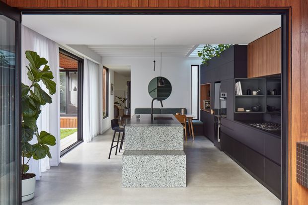 A refined open-plan kitchen and dining area viewed through a wide timber-framed opening. The room features a large speckled stone island with integrated seating, dark matte built-in cabinetry, timber overhead and wall paneling, stainless-steel appliances, concrete-look flooring, sheer curtains, and abundant glazing. A dining table and chairs sit beyond the island, with indoor planting adding a natural accent.