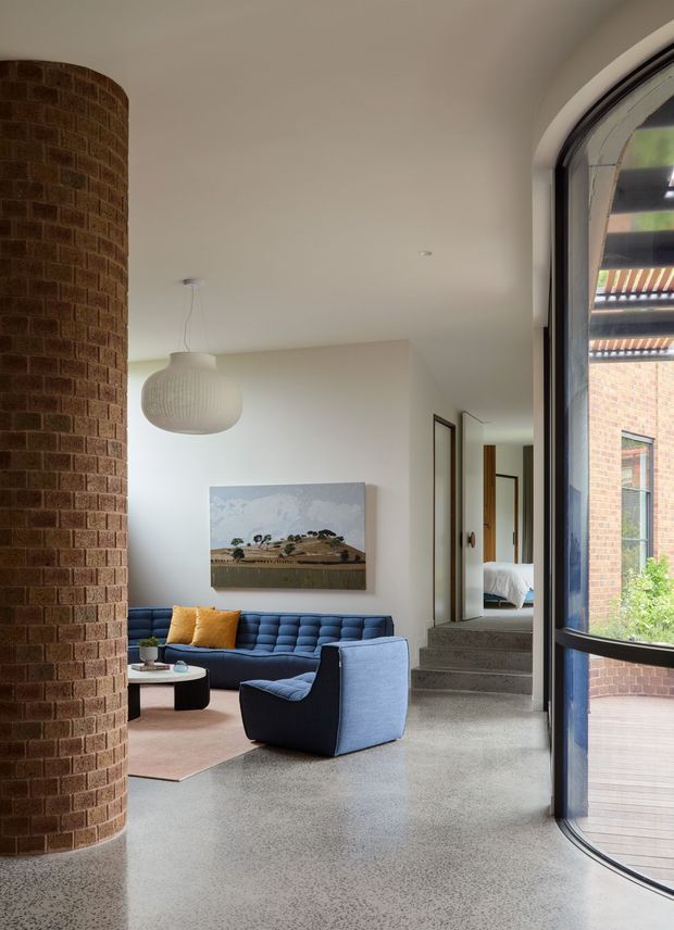 A bright contemporary living room with a curved exposed brick feature wall, polished terrazzo-like flooring, white walls, and a large arched glazed door opening to an exterior deck. The room contains a deep blue sectional sofa and matching lounge chair, a small round coffee table, a pale rug, a sculptural pendant light, and a landscape artwork. A short set of steps leads toward a bedroom visible in the background.