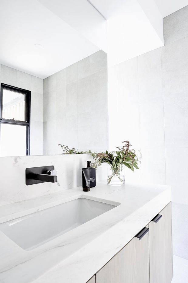 Bright minimalist bathroom vanity with a broad white stone benchtop, integrated rectangular basin, matte black wall-mounted tapware, pale large-format wall tiles, and light timber-grain cabinetry. A black-framed window and simple botanical arrangement add contrast.