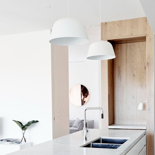 Bright contemporary kitchen interior with a pale stone island benchtop, undermount double sink and chrome gooseneck tap. White cabinetry and walls are paired with a warm timber-clad pantry or appliance wall, while two large white pendant lights hang above the island. A living area is visible beyond the kitchen, with a round wall mirror and indoor plant adding subtle decorative detail.