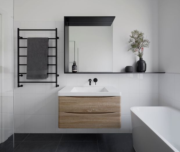 Contemporary minimalist bathroom with white tiled walls, dark charcoal floor tiles, a wall-mounted natural wood vanity with integrated white basin, matte black wall-mounted tapware, black heated towel rail, and a large black-framed mirror with a shallow shelf. A freestanding white bathtub occupies the right side, with restrained black and wood accents.