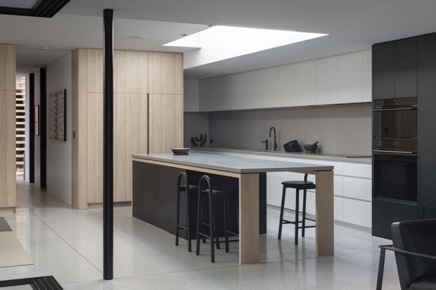 A contemporary open-plan kitchen with a large island and integrated breakfast bar. The space features pale timber cabinetry, white and dark grey joinery, a light stone-look benchtop, black appliances, and black bar stools. A large ceiling skylight provides bright natural illumination, while a hallway and part of a dark lounge chair are visible at the edges.
