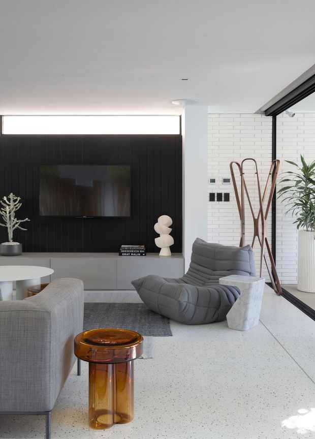 Contemporary living room with a monochrome grey, black and white scheme. A wall-mounted television is set into a black vertical-panel feature wall above low grey cabinetry, beside a white brick wall and large black-framed glazing. The room includes a sculptural grey lounge chair, grey sofa, white stone side table, amber glass occasional table, indoor plant and decorative coat stand. The space is bright, refined and minimalist with a terrazzo-like light floor.