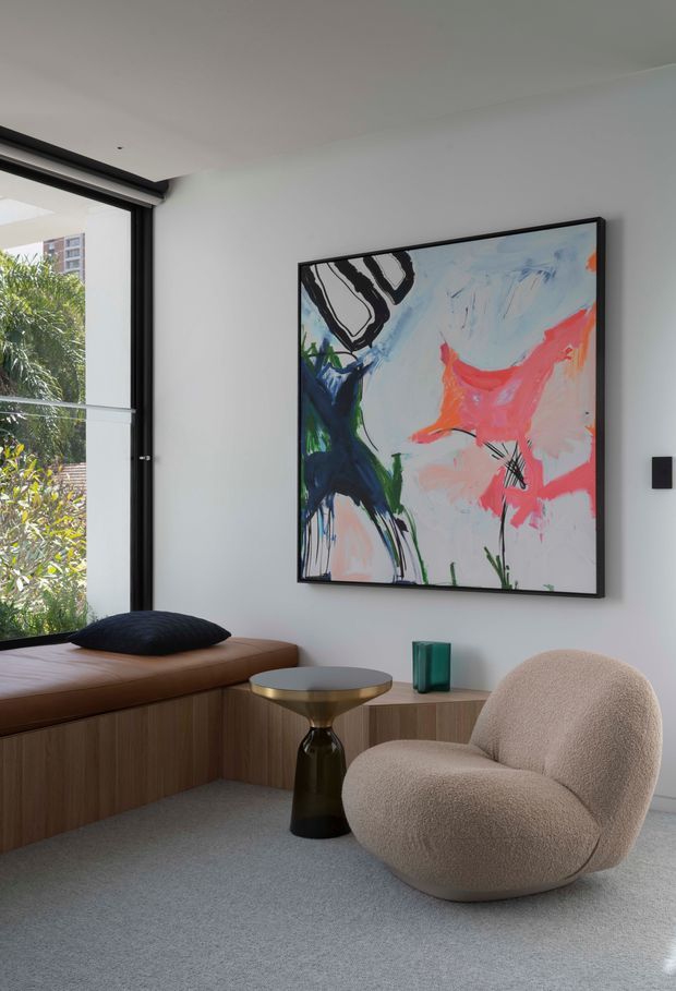 A refined contemporary living area with a floor-to-ceiling black-framed window, built-in timber window seat, sculptural upholstered lounge chair, small pedestal side table, and abstract colourful artwork on a white wall. The room has a soft grey carpet and a restrained palette accented by dark green and vivid artwork colours.